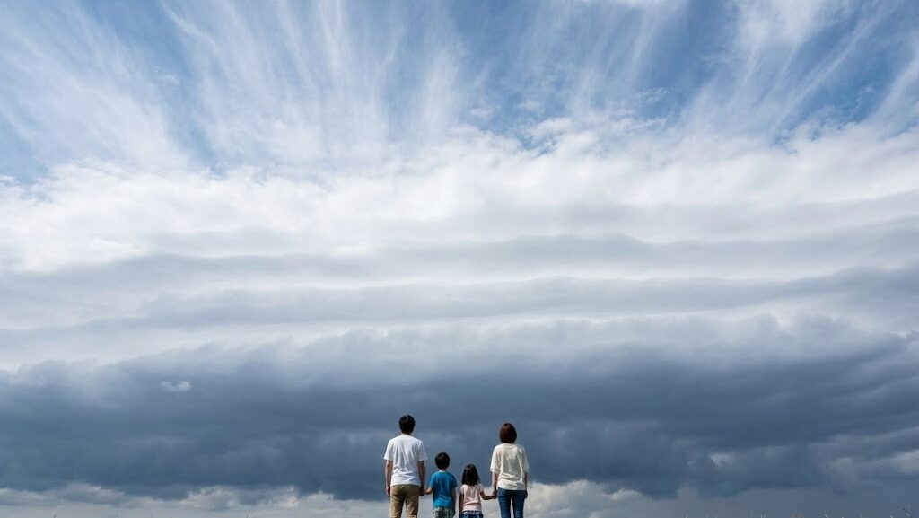 巻雲から乱層雲まで、雲の種類が奥行きで変化する空を見上げる家族の後ろ姿。台風の距離による天気変化を示す実写風画像。