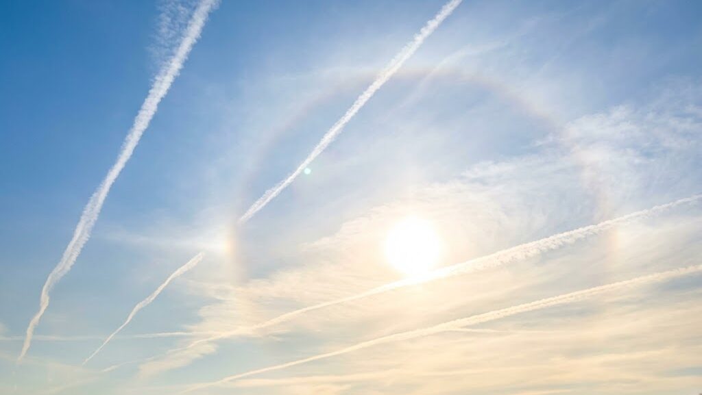 青空に長く残る飛行機雲と太陽周囲のハロ現象が見える空の写真風イメージ。台風接近の予兆を示す高層の巻層雲を表現。
