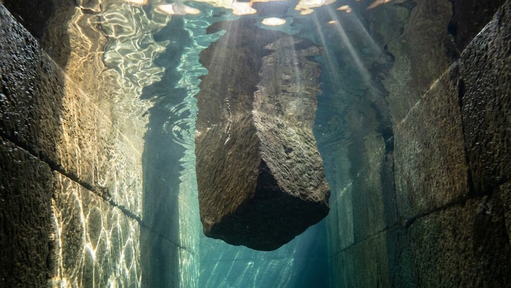 水で満たされた縦穴の中で石材が浮力で持ち上がる様子を抽象表現した実写風イメージ。