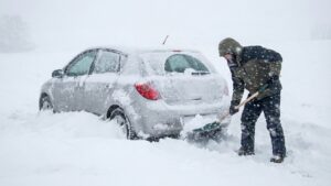 大雪で立ち往生した銀色の車の後ろで、防寒着を着た人物がスコップを使って雪をかき出している様子。