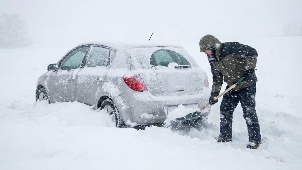 大雪で立ち往生した銀色の車の後ろで、防寒着を着た人物がスコップを使って雪をかき出している様子。