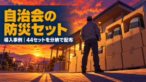 夕焼けの集会所の前に積まれた防災リュックの箱と、見上げる町内会役員のイメージ